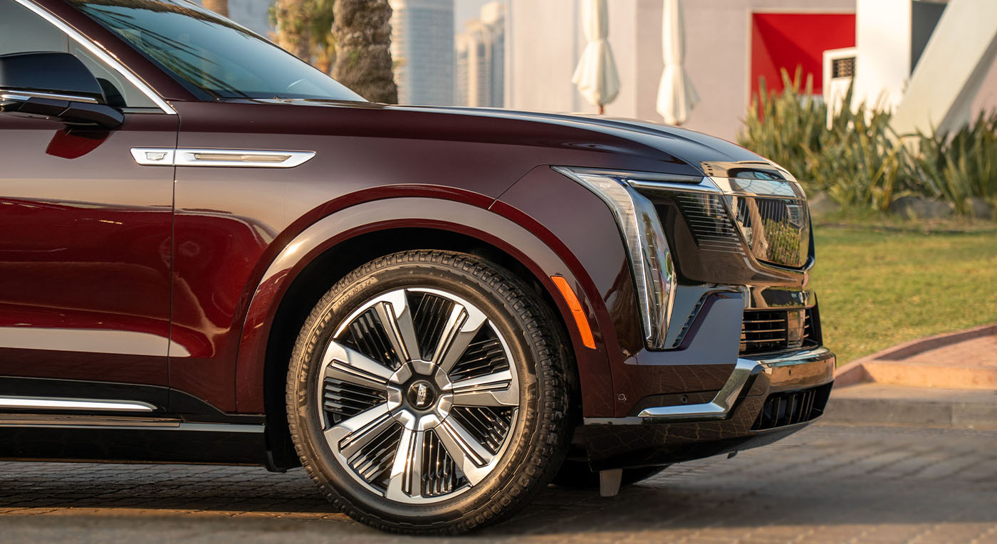 Close-up of a dark red Cadillac Escalade IQ luxury SUV showcasing the front wheel, chrome accents, and sleek design, parked in an urban setting with greenery.